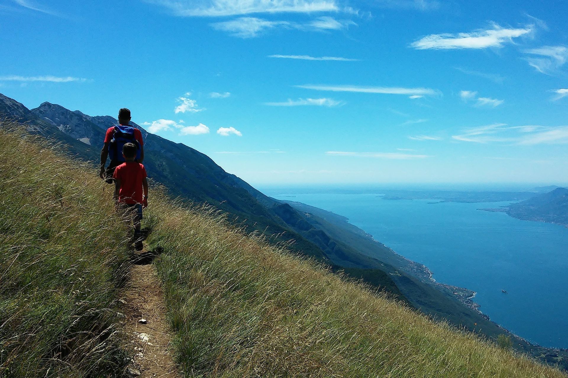 Due escursionisti su uno stretto sentiero di montagna con vista su un oceano blu e colline in lontananza.