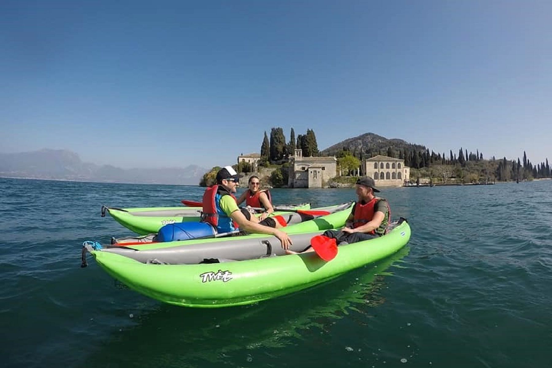 Persone in kayak su un lago vicino alla riva di un castello, sotto un cielo azzurro e limpido.