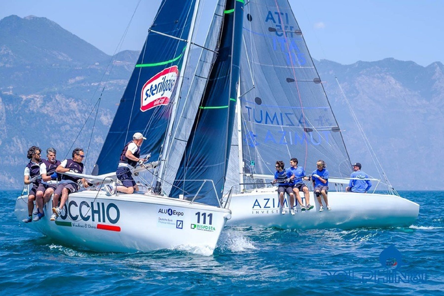 Due barche a vela che gareggiano su acque blu, con gli equipaggi in equilibrio sui ponti e le montagne sullo sfondo.
