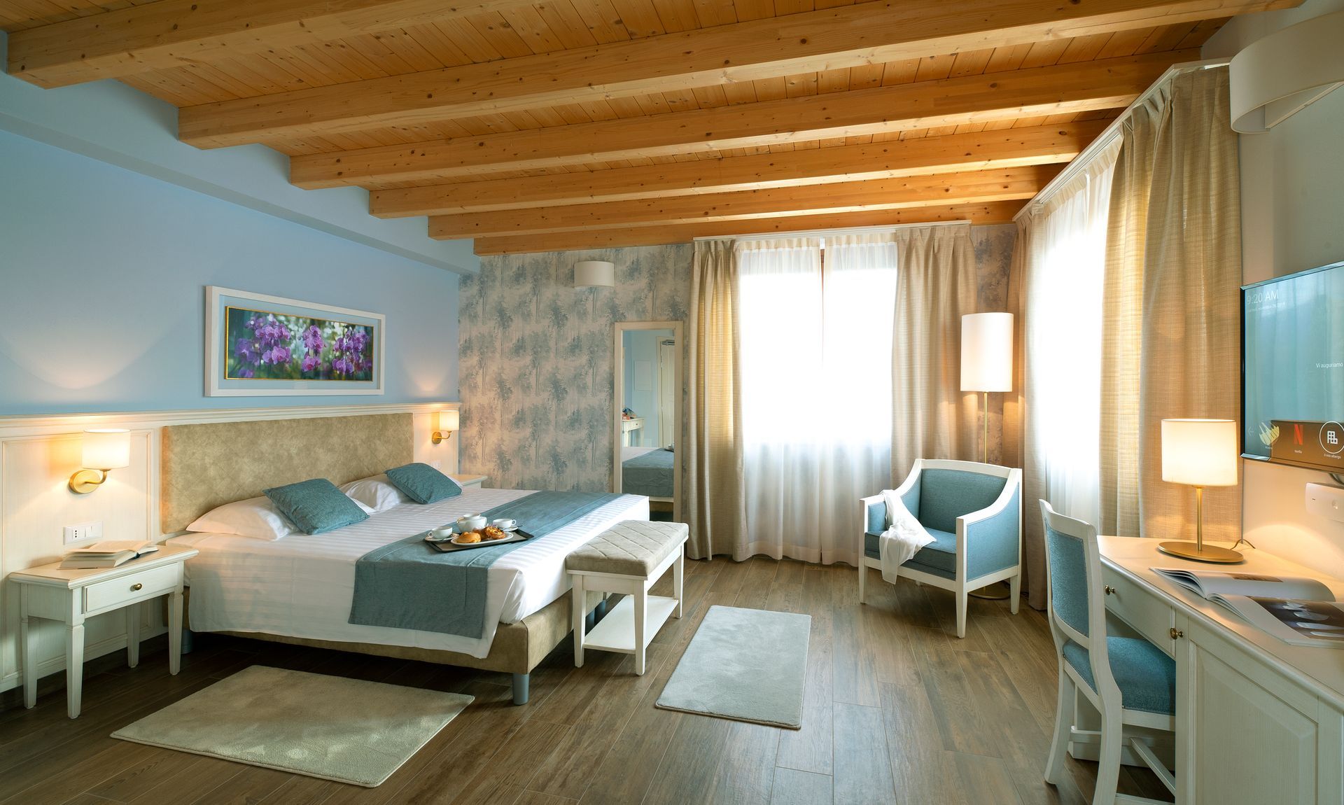 Gemütliches Schlafzimmer mit Holzbalkendecke, großem Bett, Sessel, Schreibtisch und hellen Vorhängen.