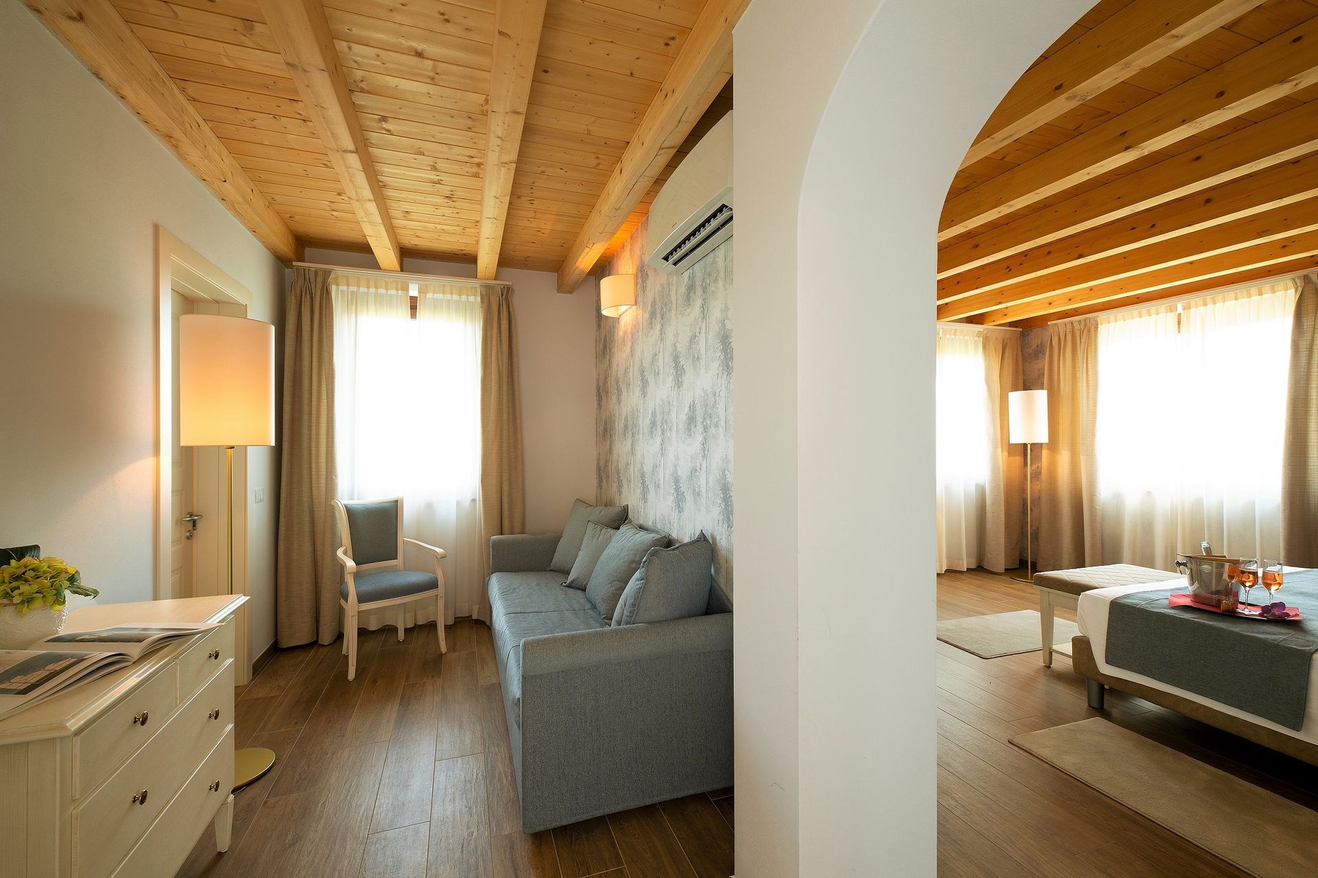 Modernes Wohn- und Schlafzimmer mit Holzdecke, weißen Säulen, grauem Sofa und warmem Fensterlicht