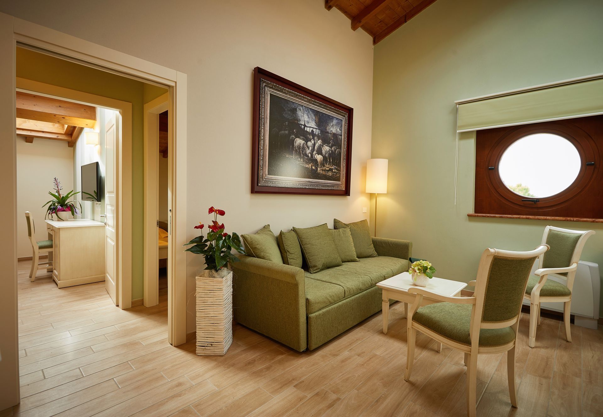 Helles Wohnzimmer mit grünem Sofa, Sesseln, Stehlampe und Zugang zu einem Badezimmer, mit Wandgemälde und rundem Fenster.
