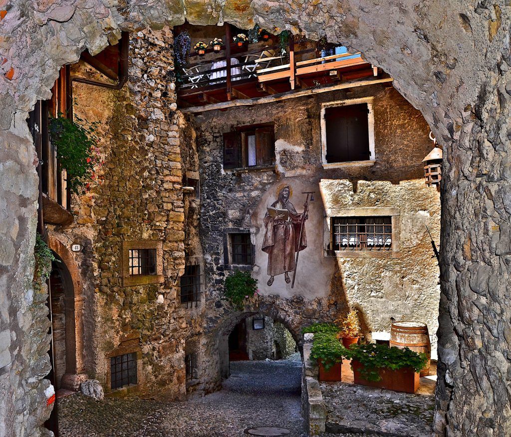 Un arco in pietra incornicia un antico cortile rustico con muri consumati dal tempo, finestre e piante in vaso.