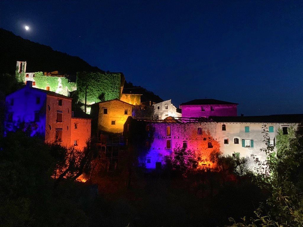 Villaggio collinare illuminato da luci colorate di notte, con edifici rischiarati contro un cielo scuro.