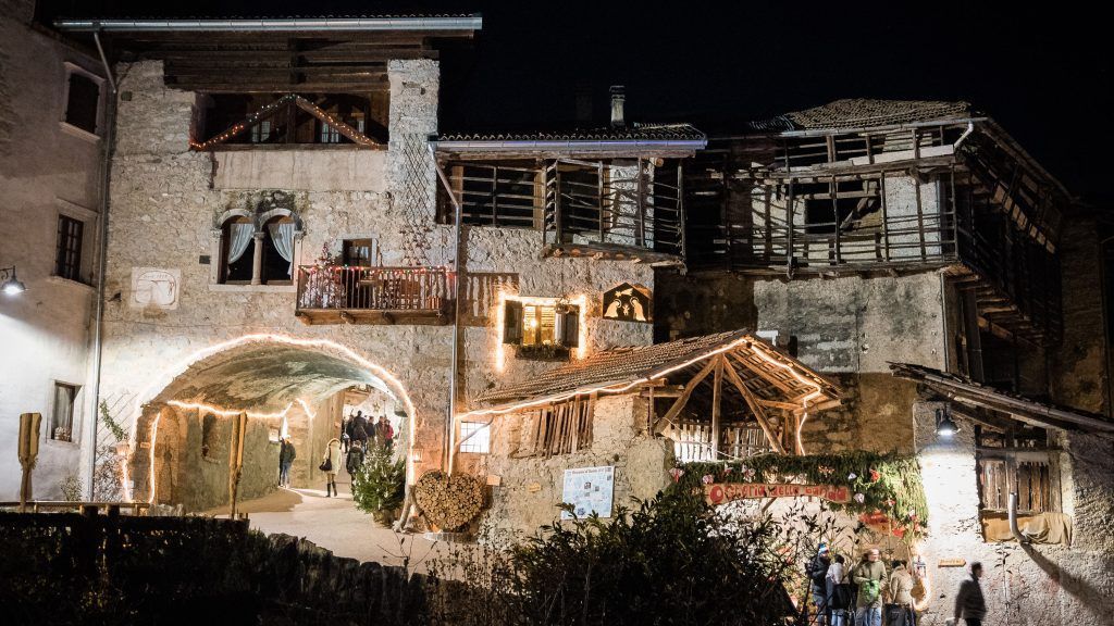 Veduta notturna di un rustico edificio in pietra con arco illuminato, balconi e terreno innevato.