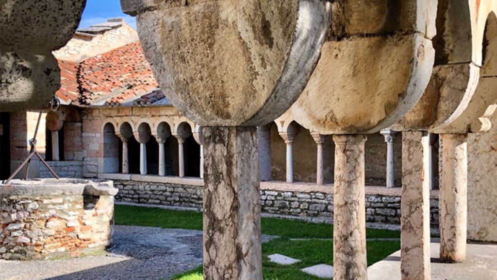Cortile in pietra con passaggio ad arco, tetto di tegole rosse e grande scultura rotonda sovrastante.
