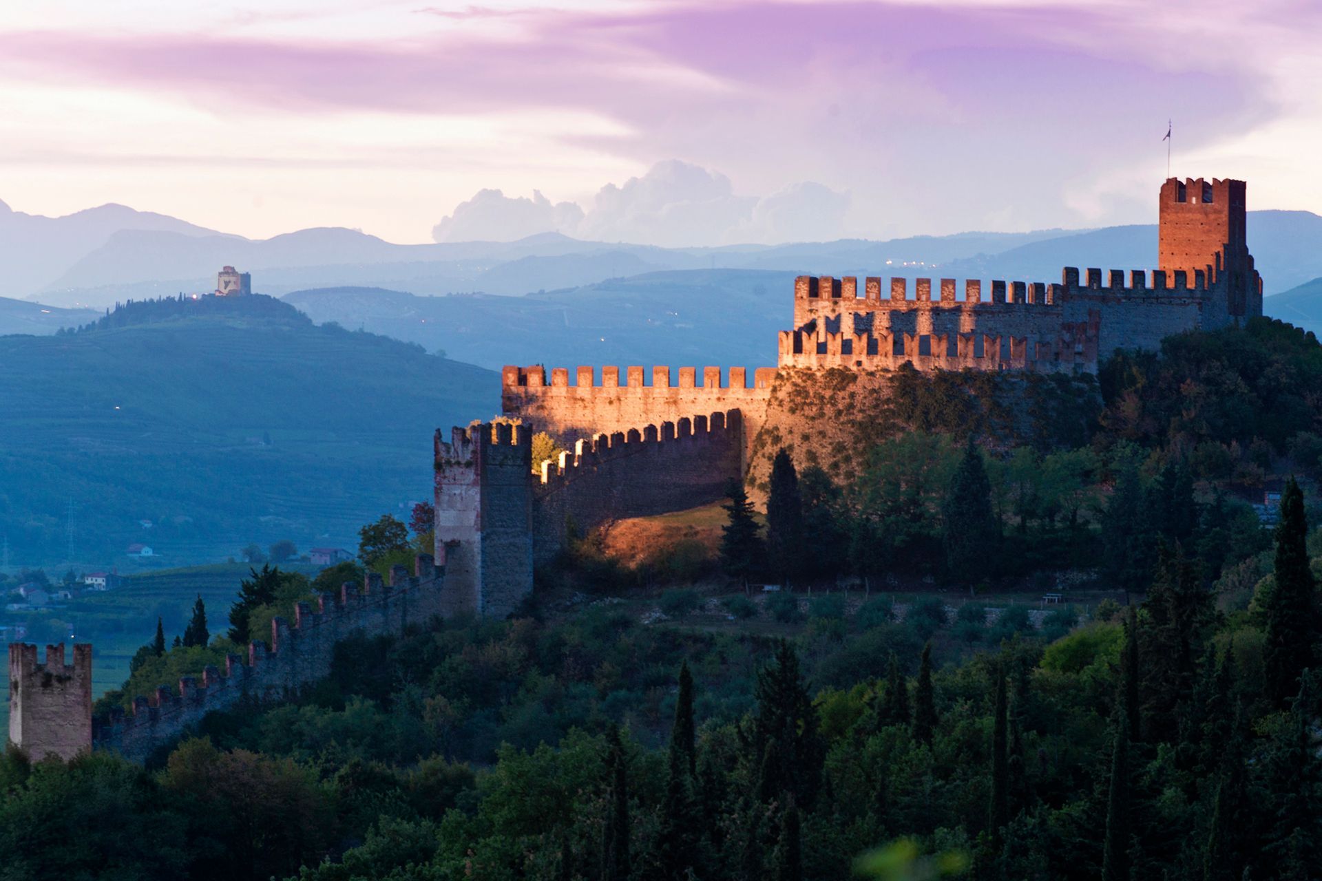 Fortezza di pietra illuminata dal sole su una collina al crepuscolo, con vista su montagne blu nebbiose sotto un cielo rosa