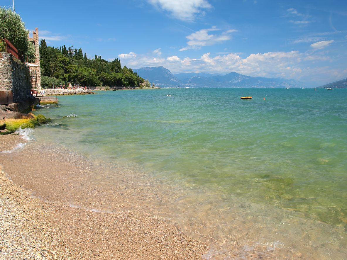 Spiaggia soleggiata di ciottoli con acqua cristallina turchese, montagne in lontananza e un cielo azzurro.