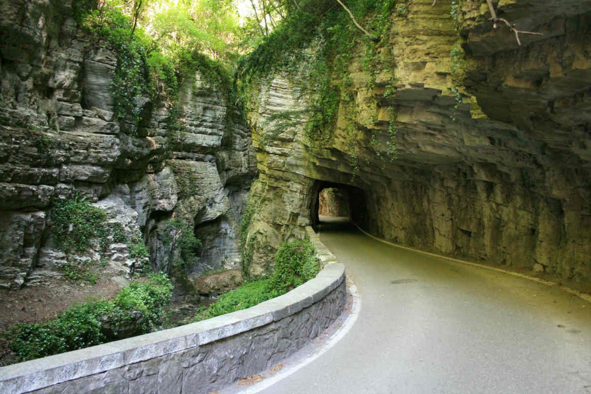 Strada tortuosa che attraversa una galleria rocciosa a fianco di una scogliera verde e di un muro di pietra.