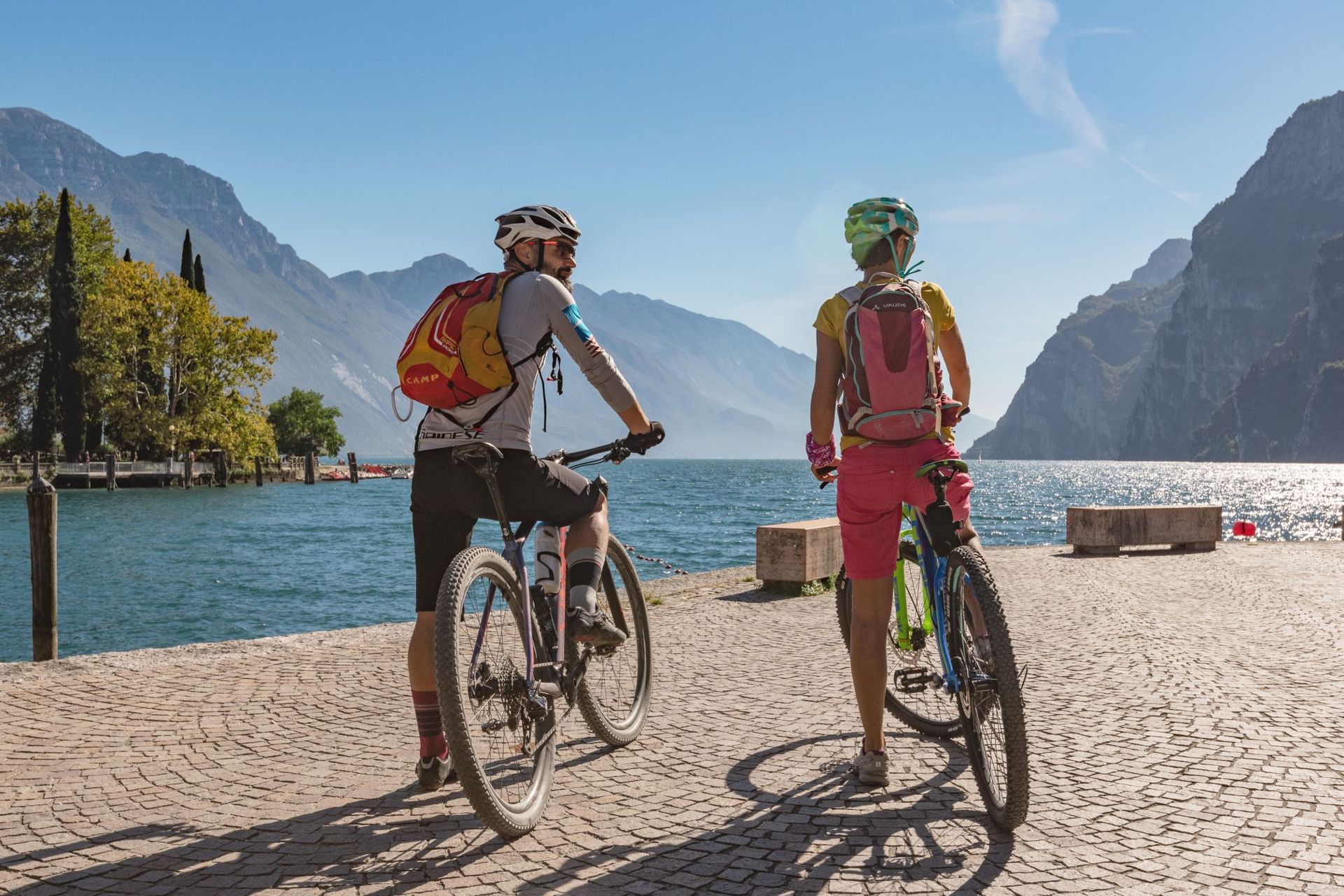 Due ciclisti con gli zaini si fermano su una spiaggia in riva a un lago, con le montagne sullo sfondo.
