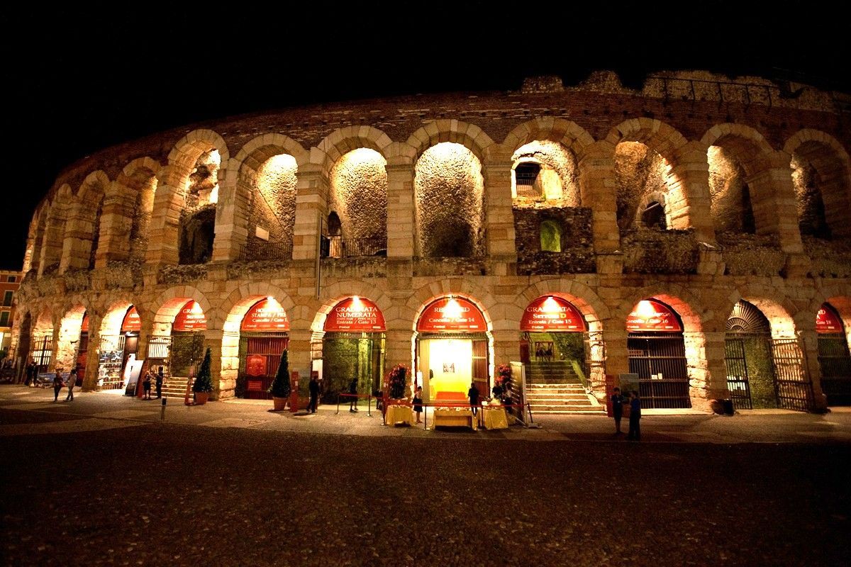Anfiteatro in pietra illuminato di notte, con aperture ad arco e persone radunate all'ingresso.