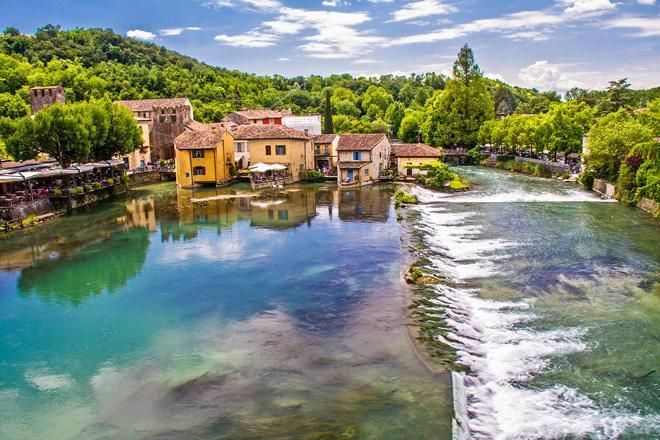 Un pittoresco villaggio fluviale con case in pietra, acqua azzurra e una piccola cascata sotto un cielo luminoso.