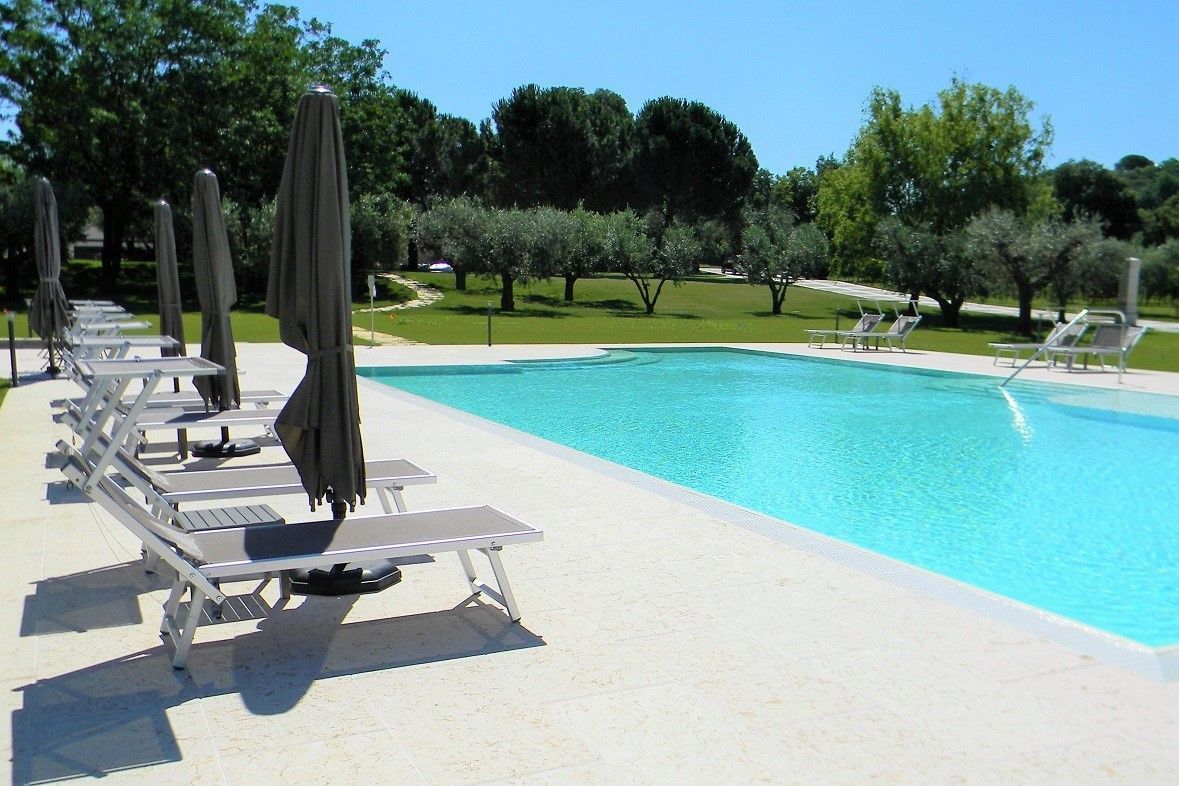 Piscina all'aperto con lettini, ombrelloni chiusi e alberi in una giornata di sole.