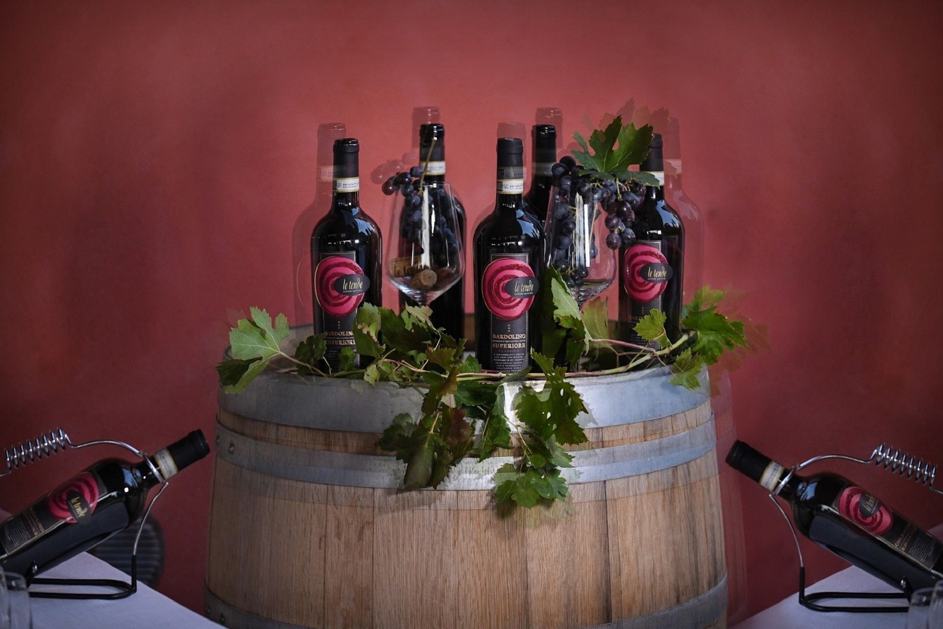 Bottiglie di vino rosso disposte in un'esposizione rustica a forma di botte, con foglie verdi su sfondo rosso.