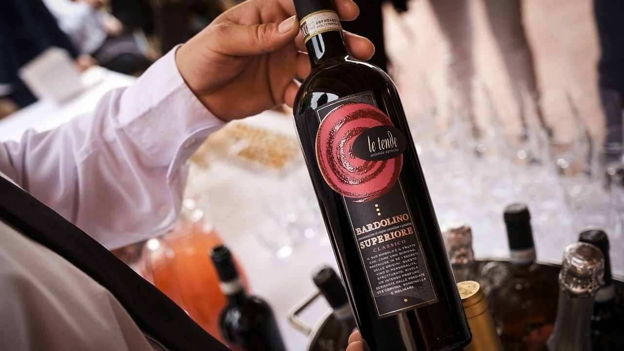 Mano che regge una bottiglia di vino scuro con etichetta rossa durante una degustazione.