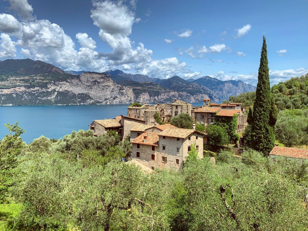 Villaggio lacustre con case in pietra, cipressi e montagne sotto un cielo azzurro brillante