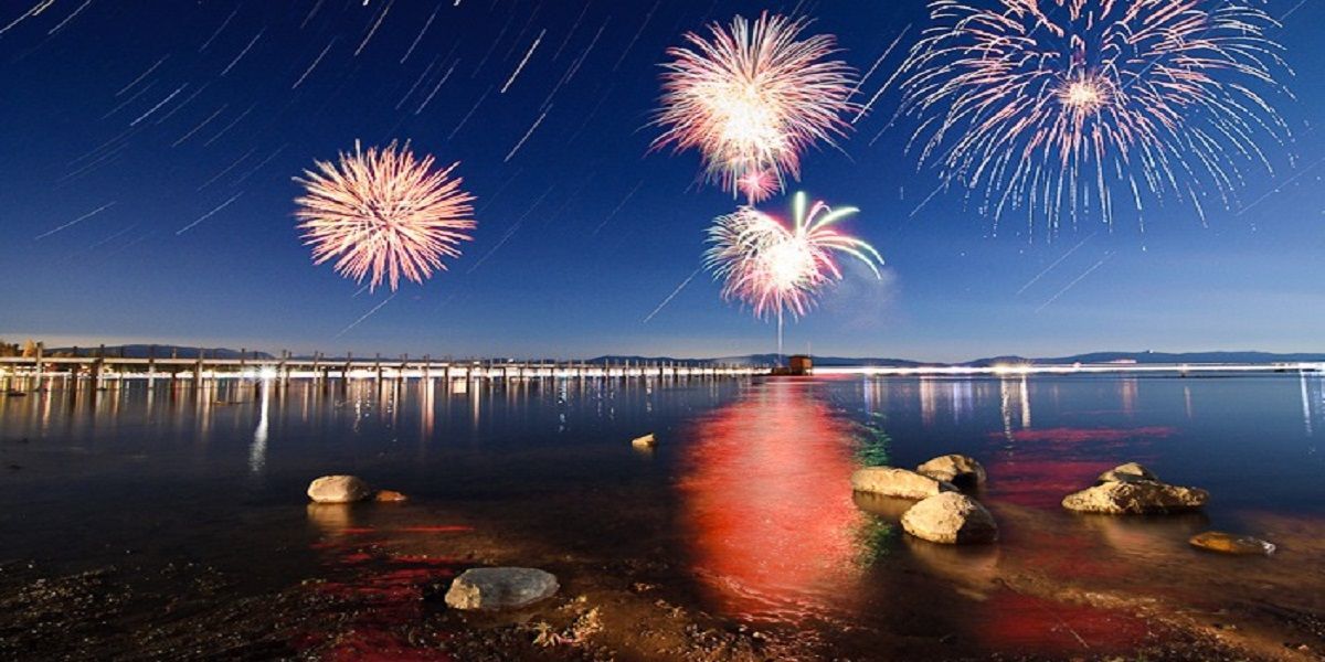 Fuochi d'artificio su un tranquillo litorale di notte, che si riflettono sull'acqua vicino a scogli e a una riva illuminata.