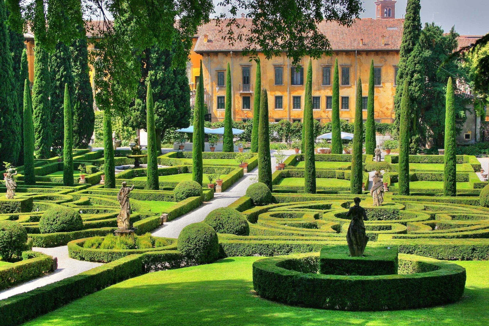 Giardino formale con siepi ben curate, viali alberati e una grande villa sullo sfondo.