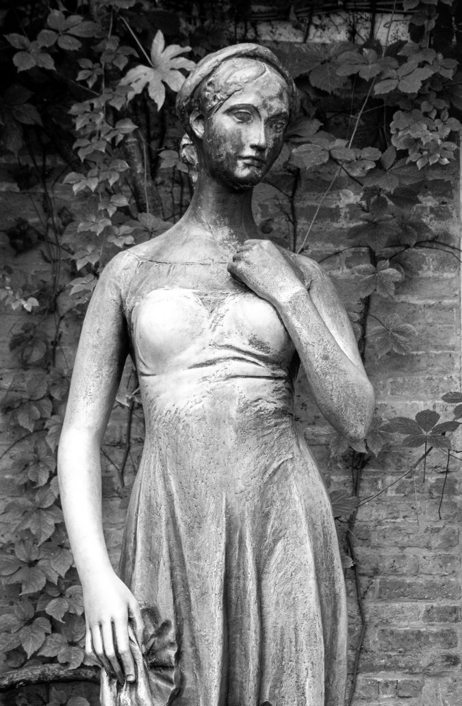 Statua in bianco e nero di una donna in abito, in piedi contro un muro di mattoni con vegetazione alle sue spalle.
