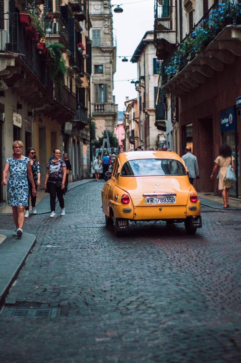 Un'auto d'epoca gialla percorre una strada acciottolata fiancheggiata da vecchi edifici e pedoni.