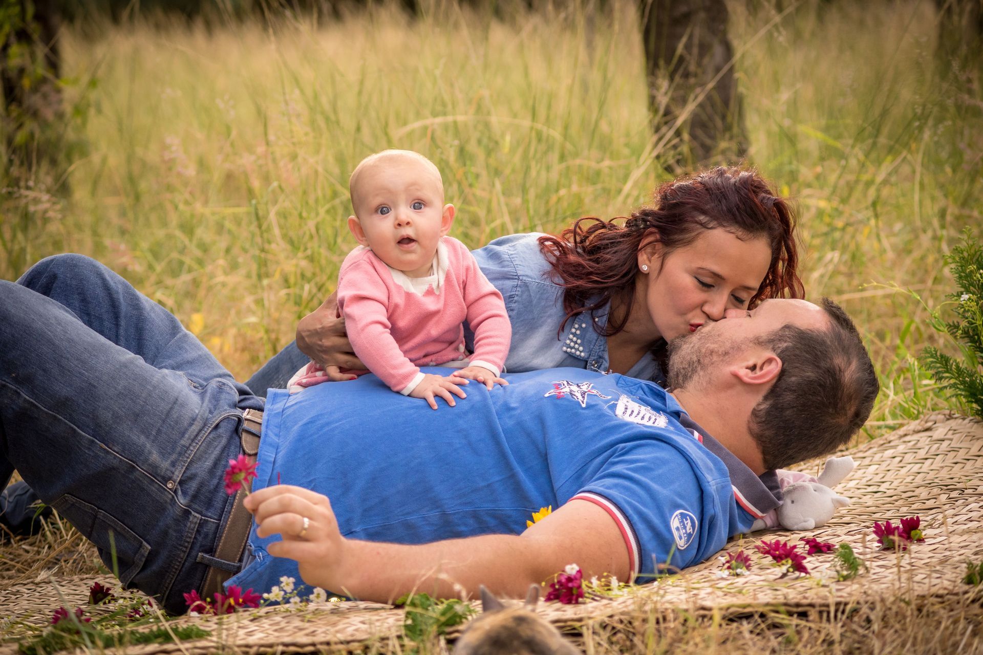 Woman and man lying in grass with a baby between them, outdoors on a blanket, smiling and cuddling