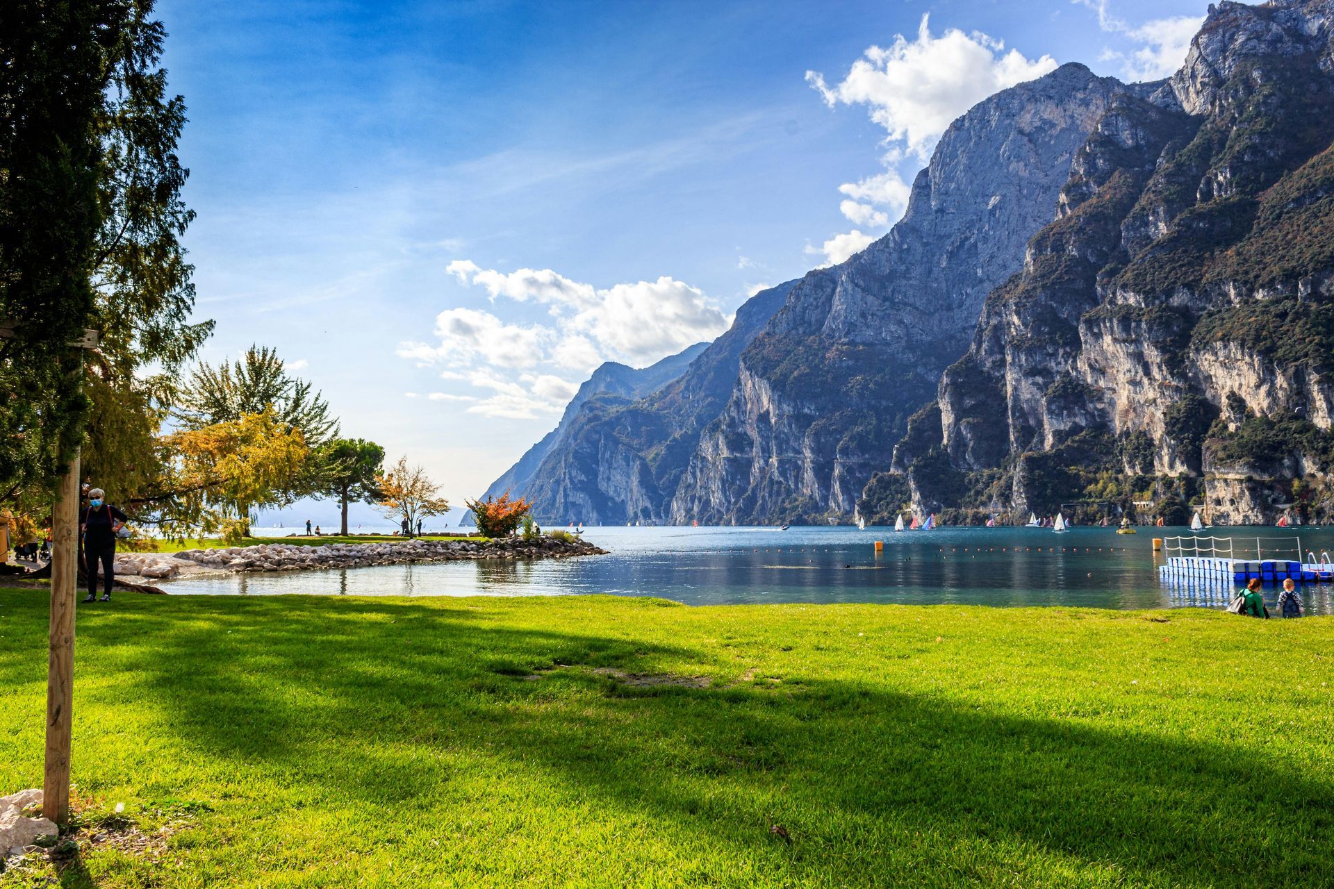 Parco in riva al lago con prato verde, acque blu calme e montagne aspre sotto un cielo luminoso
