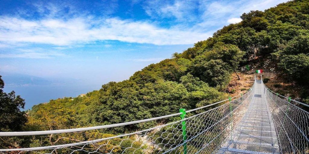 Un sentiero su un ponte sospeso attraversa una verde collina sotto un cielo azzurro, con una persona in lontananza.