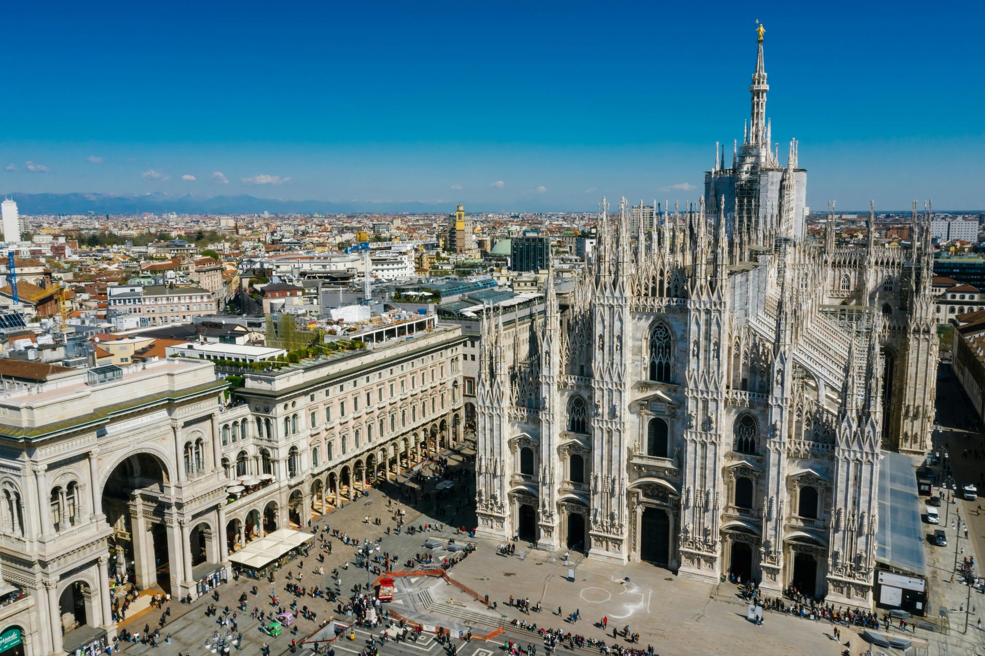 Veduta aerea del Duomo di Milano e di Piazza del Duomo sotto un cielo azzurro e limpido.