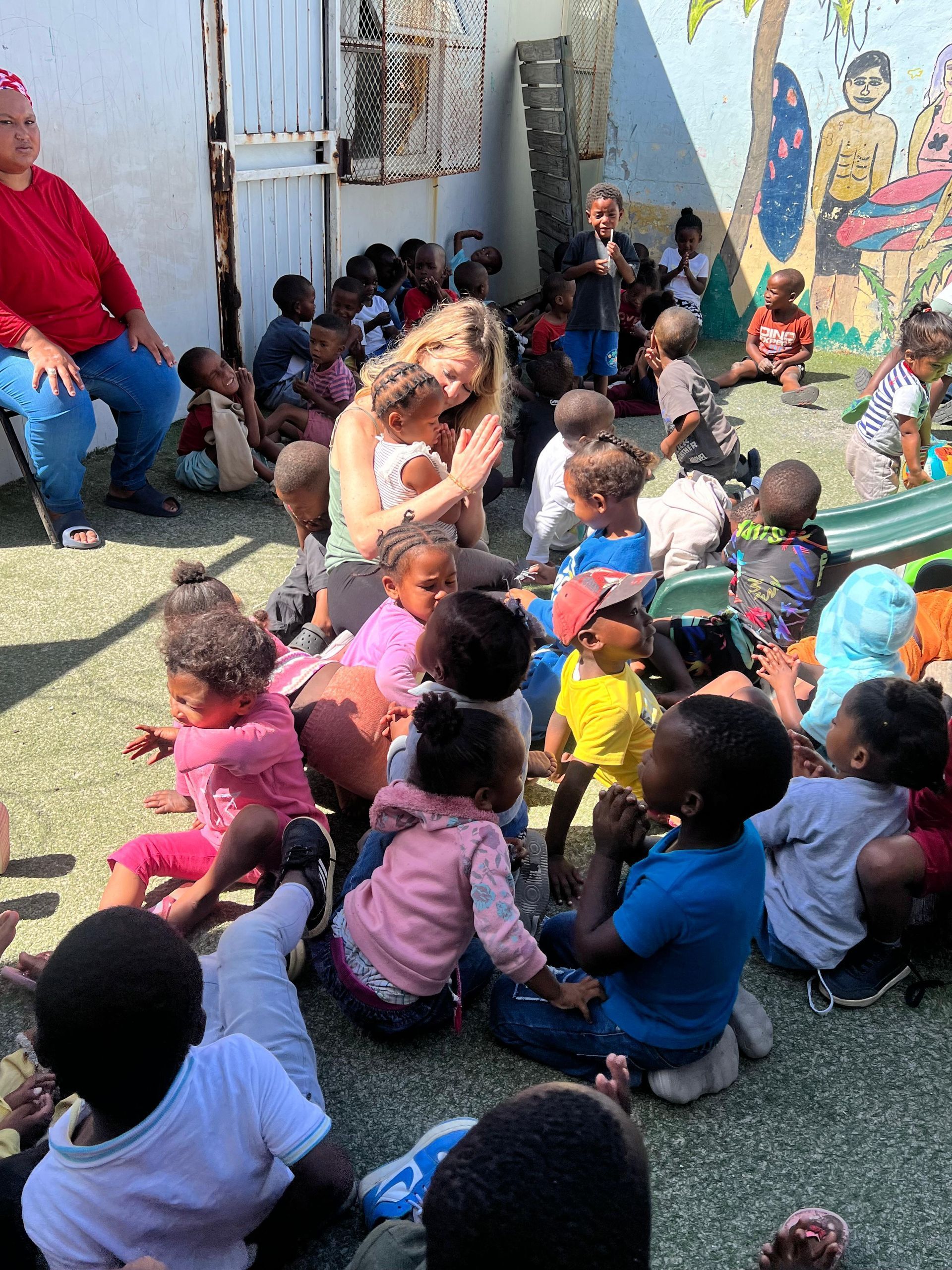 A woman is talking to a group of children sitting on the ground.
