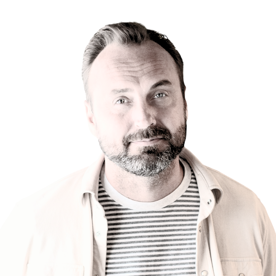 Man met baard, in gestreept shirt en open beige overhemd, kijkt in de camera.