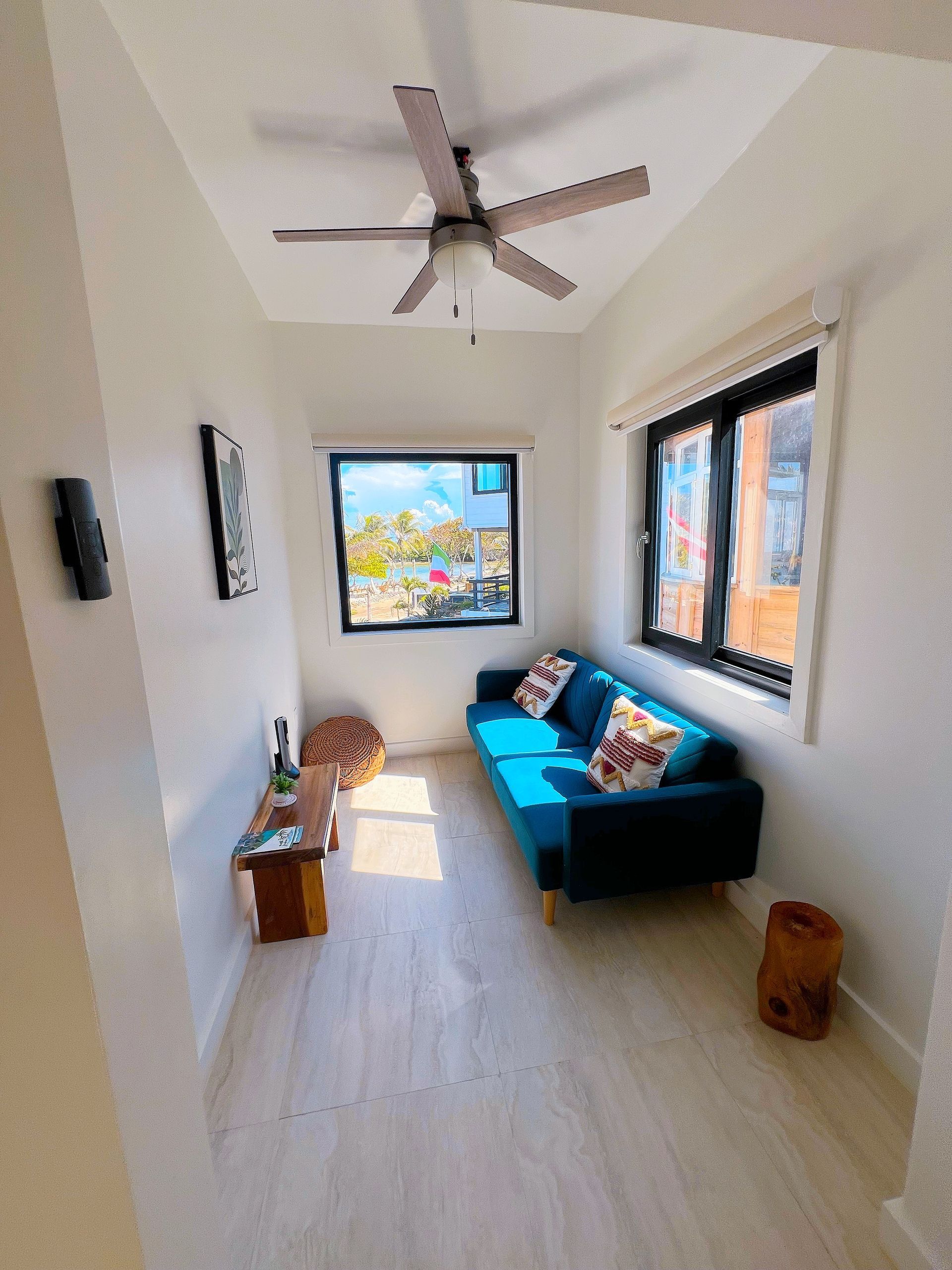 Living room with blue sofa, wooden table, and large windows, white walls, and a ceiling fan.