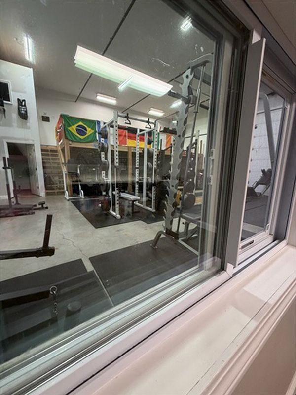 Gym interior seen through a window, with weight racks, mirrors, and a Brazilian flag overhead.