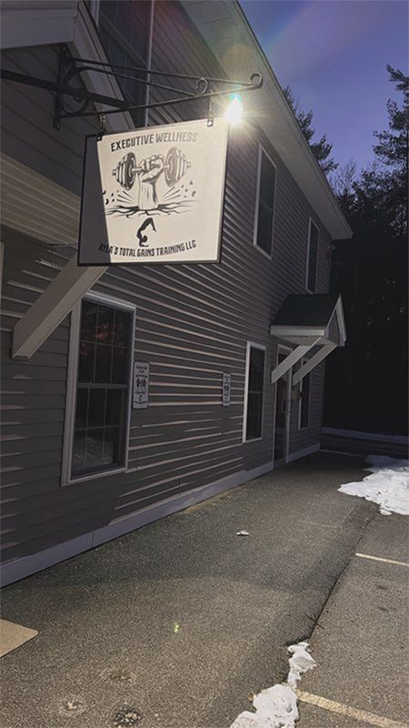 Building entrance at dusk with illuminated hanging sign, gray siding, and a snow-covered walkway.