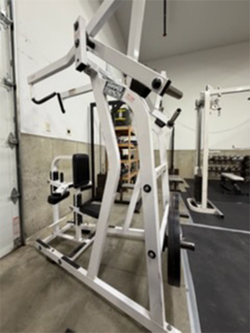 White plate-loaded gym machine in an empty fitness room with black padding and mirrors.