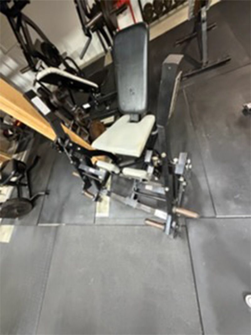 Weight machine with black frame and white seat in a gym on dark tile flooring.