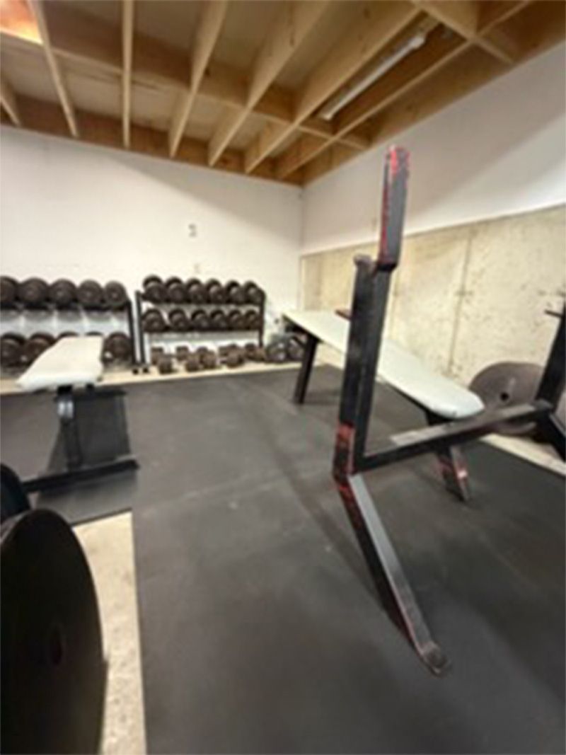 Small home gym with weight racks, bench, squat stand, and black mat flooring under exposed beams
