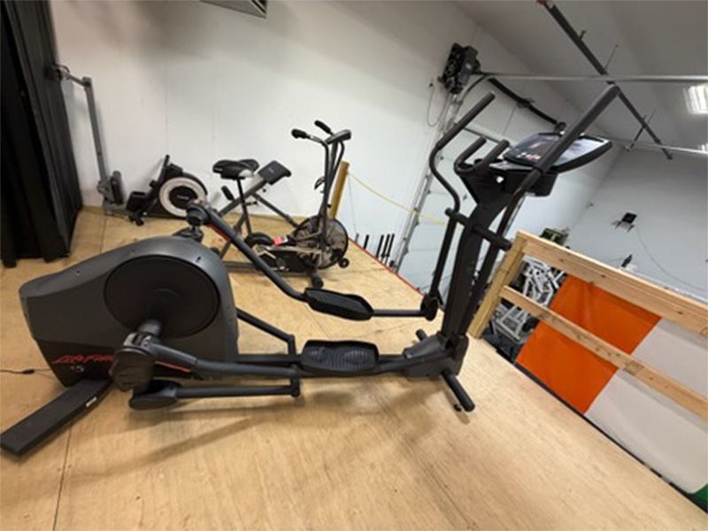 Gym with two stationary exercise bikes on a wooden floor, one lying on its side near a loft railing.