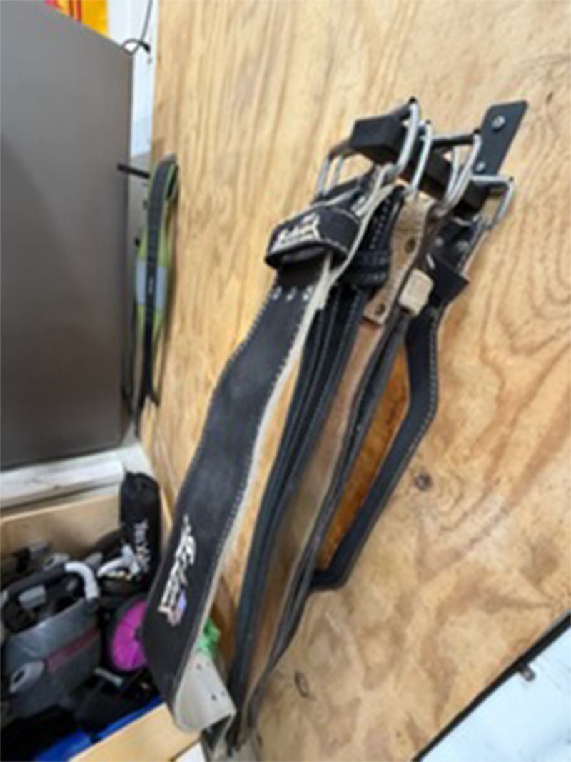 Black straps with silver buckles hanging on a plywood wall beside a shelf of gear.