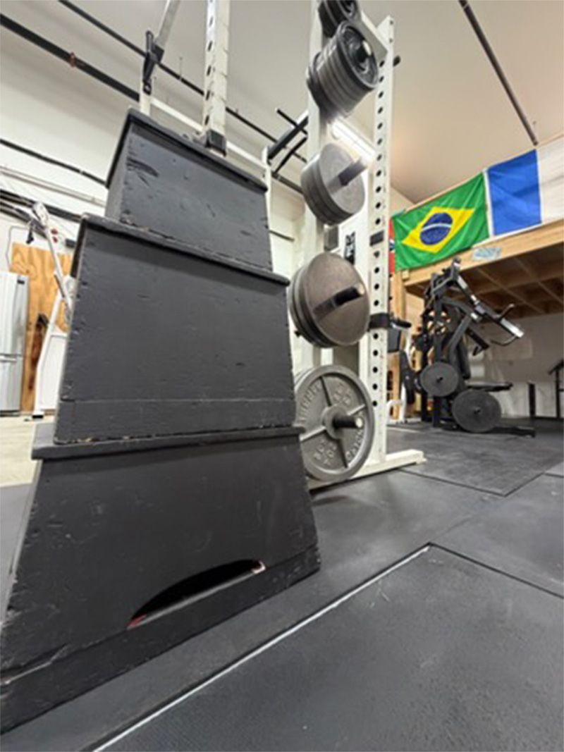 Indoor gym equipment with stacked black platforms, metal apparatus, and a Brazilian flag in the background
