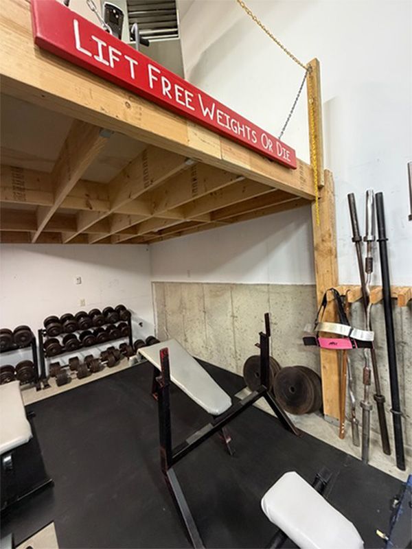 Home gym corner with a lofted wooden platform, bench press, dumbbells, and barbells stored on the wall