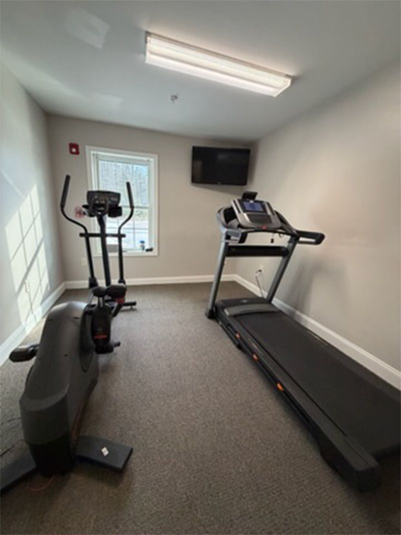 Small home gym with a treadmill, exercise bike, wall-mounted TV, and overhead lights.