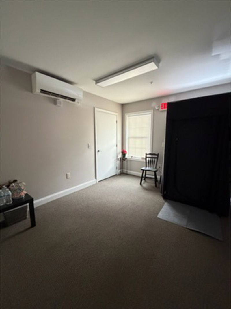 Empty room with beige carpet, white walls, ceiling lights, wall AC, and a black curtain by a chair.