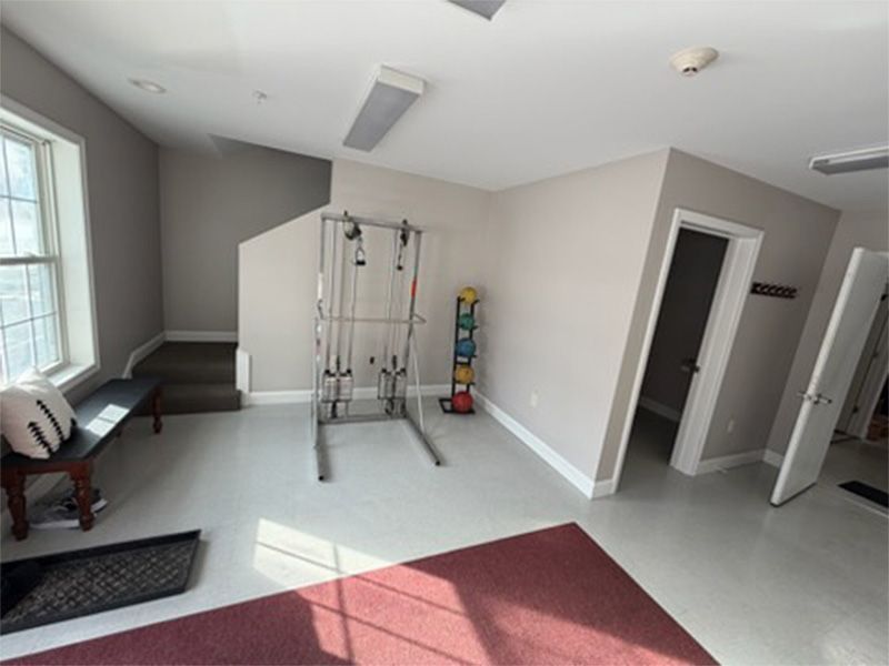 Bright home gym with cable machine, wall mirror, bench, stairs, and white walls with red carpet.