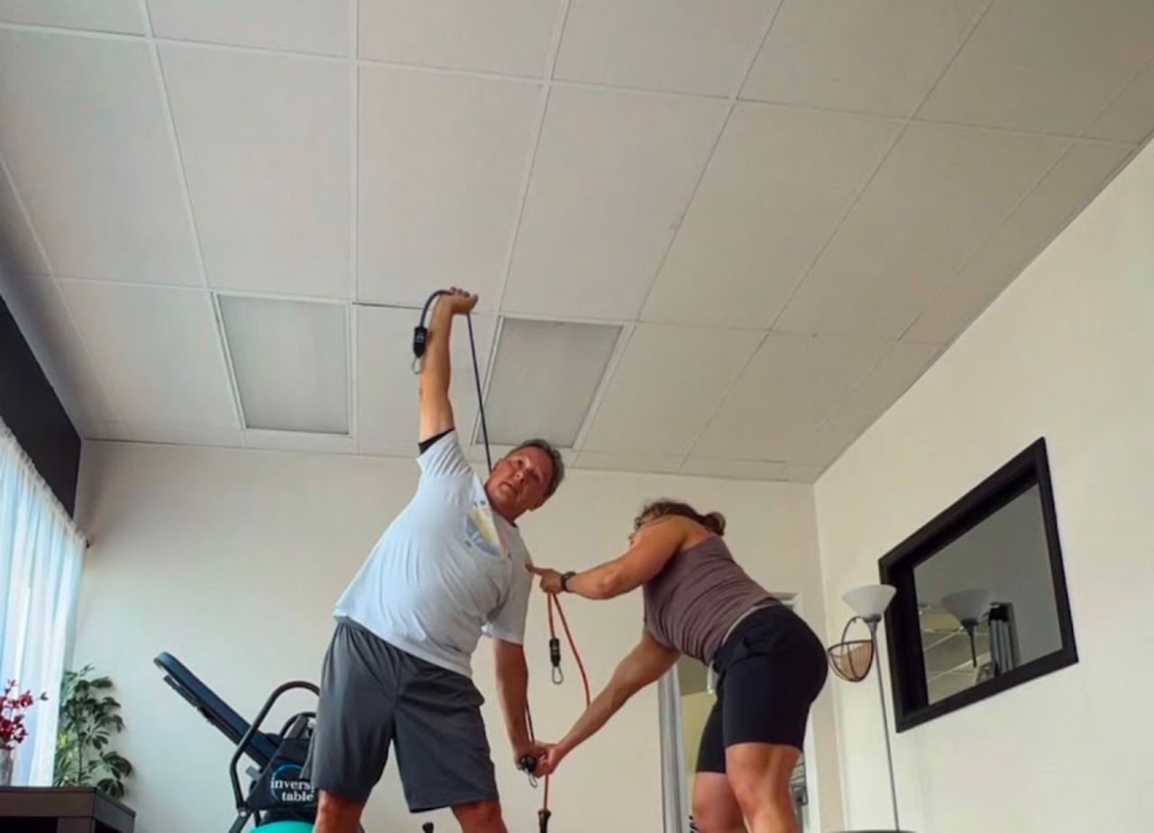 A man stretches with resistance bands as a woman assists him in a room with exercise equipment.