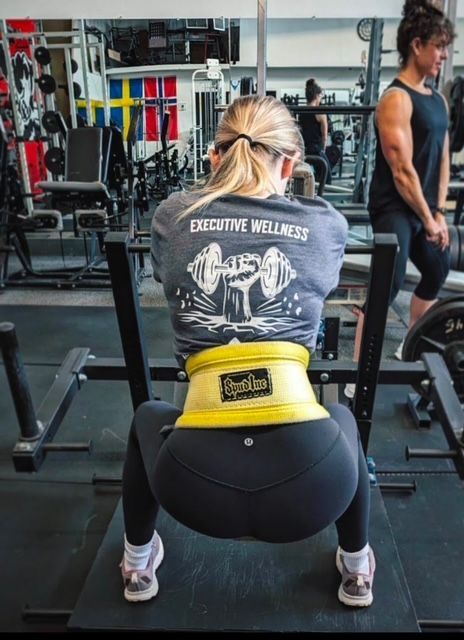 Woman squatting in a gym, wearing a yellow weightlifting belt. Another woman watches in the background.