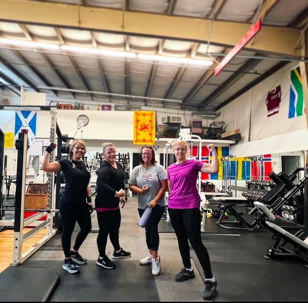 Four women flex in a gym. Weights and equipment visible. They smile, wearing athletic clothing.