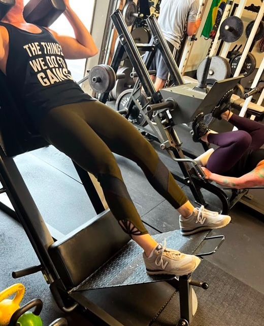 Person exercising on a leg press machine at a gym. Dark leggings, black top, and sneakers.