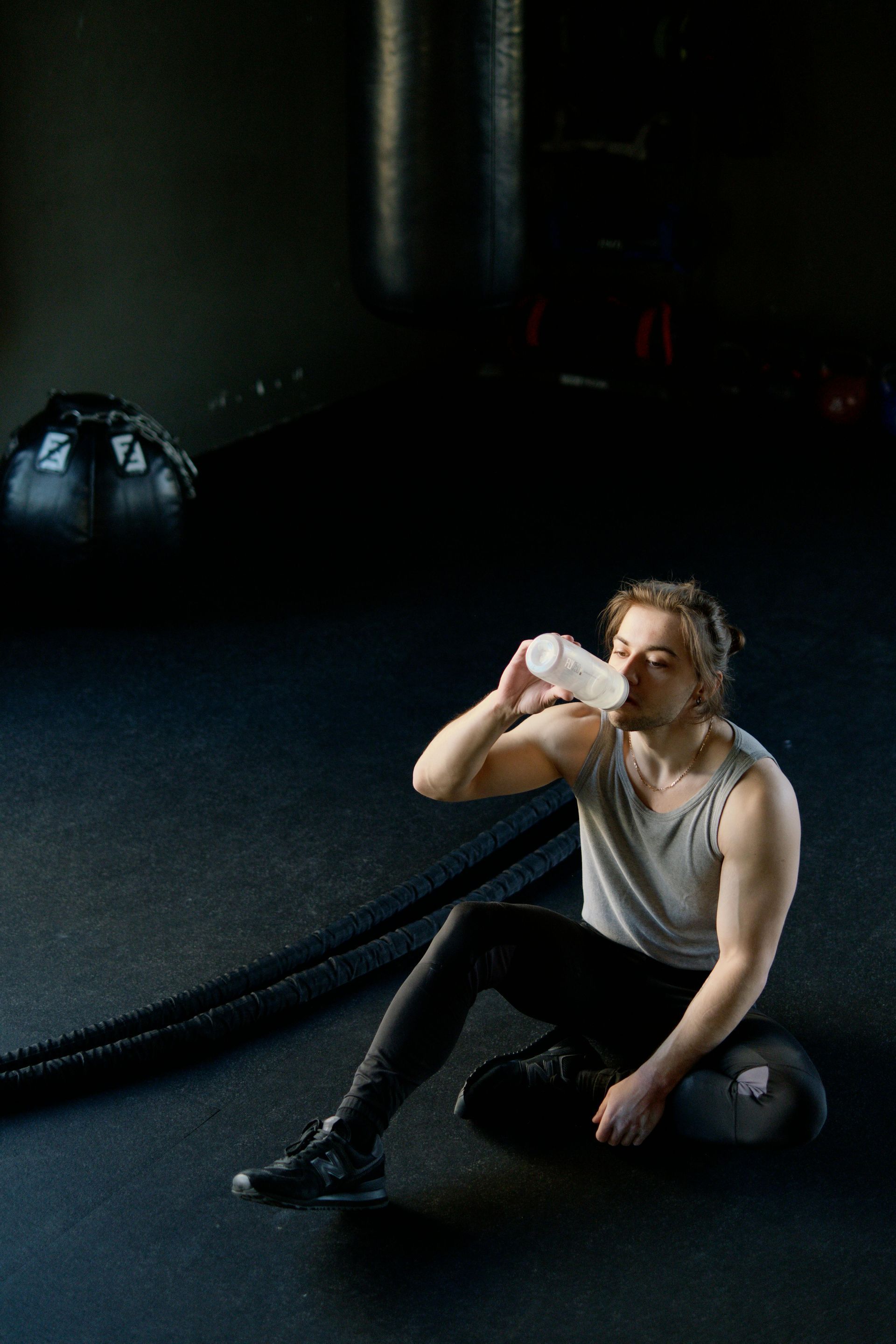 Person drinks water, resting on a medicine ball in a gym. Black floor, battle rope, and bag in background.