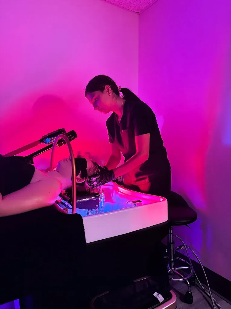 Woman in black uniform tending to a client's face illuminated by pink and blue lights in a salon.