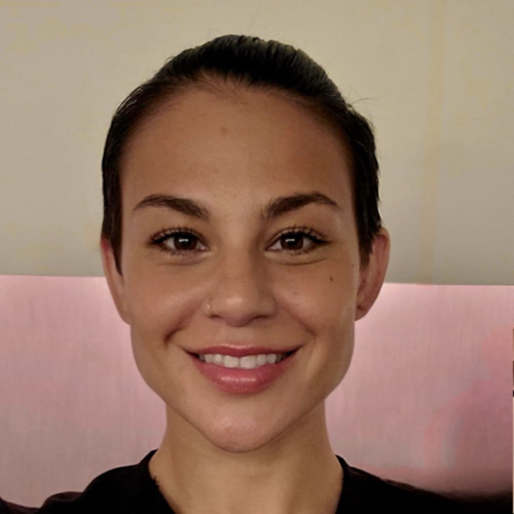 Woman with short dark hair smiles, sitting in front of a pink couch.